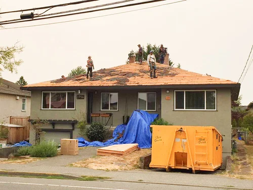 Roofing crew working on a stripped roof with an orange debris dumpster outside